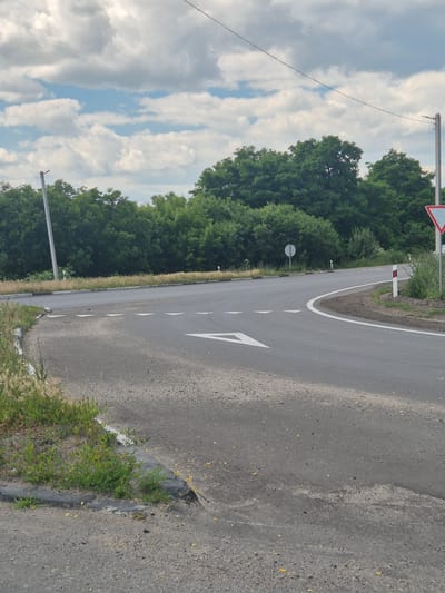 The image shows an outdoor scene at a road intersection. The road surface is asphalt and appears somewhat worn. White directional arrows are painted on the road, indicating a turn. A white line marks the edge of the road. Along the side of the road, there is dry grass and some sparse green vegetation growing near a concrete curb. In the background, a dense line of green trees forms a natural barrier. Overhead, a cloudy sky with patches of blue is visible, with a power line stretching across the frame. There are also several utility poles and road signs present, including a triangular warning sign and a round white sign. The scene is set during the daytime. The location is identified as Pidhaitsi, Ukraine.