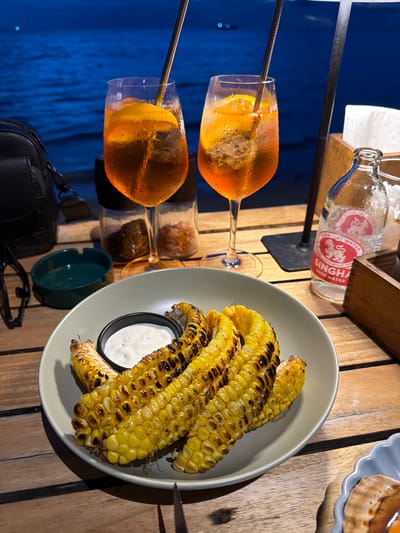 Two glasses with orange beverages and ice, each with a metal straw and a slice of orange, sit on a wooden table. A bottle of Singha Soda Water is visible behind the glasses. A plate of grilled corn on the cob with a small bowl of white dip is also on the table. In the background, a dark blue surface suggests water, and a black leather bag rests on the table. The scene appears to be outdoors at dusk or night, with artificial lighting illuminating the food and drinks. The location is identified as Ko Tao Subdistrict, Thailand. No people are visible in the image.