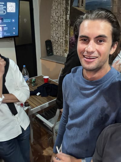 A candid, close-up image shows a man smiling into the camera, seated with others at an outdoor table. The man is wearing a blue sweater and holds a cigarette. Part of another person is visible, wearing a white shirt and black pants. A TV screen is in the background, along with a stone-textured wall. On the wooden table are several items including a red cup, a green can, a bottle of water, and what appears to be a brown liquid in a glass or container. The location is San Pedro Cholula, Mexico.