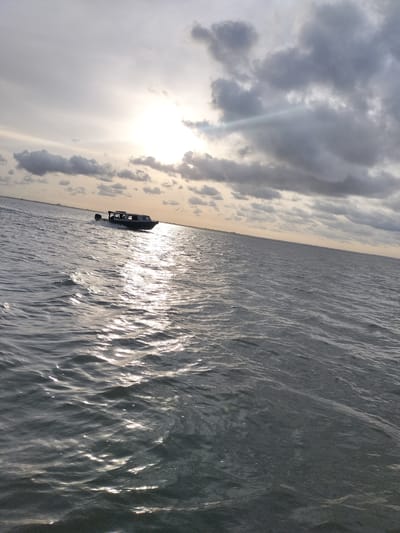 A low-angle, eye-level shot captures a black and white motorboat with passengers on the right side, cruising through the dark, choppy waters of a sea. The boat is angled towards the left of the frame, with its dark hull and white cabin visible, and its outboard motor can be seen at the stern. The sun is positioned in the upper left quadrant of the image, partially obscured by dark, dramatic clouds, casting a shimmering reflection across the water's surface. The horizon line is visible in the distance, with a faint outline of a city skyline, possibly indicating the location of Ikoyi, Nigeria. The overall lighting is dim, with the sun's rays piercing through the clouds, creating a sense of dynamic weather conditions.