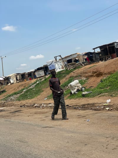 The image depicts a street scene in Kwali, Nigeria. An individual stands on the right edge of a paved road, wearing a dark shirt and patterned pants. Along the left side of the road is an area characterized by a mix of vegetation and trash, adjacent to a series of makeshift dwellings or shacks constructed from various materials. Power lines are visible against a blue sky with scattered clouds.