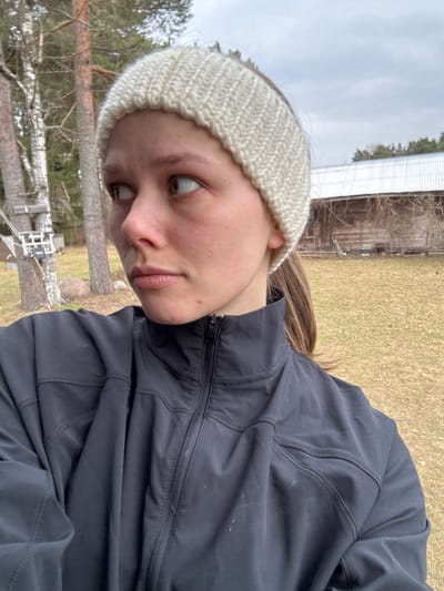 This image depicts a young female outdoors in Dubki, Russia. She is positioned in a medium shot, angled slightly towards the viewer's left, with her head turned and gaze directed to the viewer's right. She wears an off-white knitted headband with a chevron pattern and a dark-colored, zippered jacket. Brown hair is visible below the headband and jacket collar. Her facial expression is neutral. The background features a rural or semi-rural environment with dry, yellowish-brown grass covering the ground. Multiple trees are present, including two prominent light-barked trees resembling birch, and several darker-barked trees. A white, rectangular object is attached to one of the trees on the left. In the mid-ground on the right, a weathered wooden building with a corrugated metal roof is visible, partially obscured by bare vines or branches. The sky is overcast.