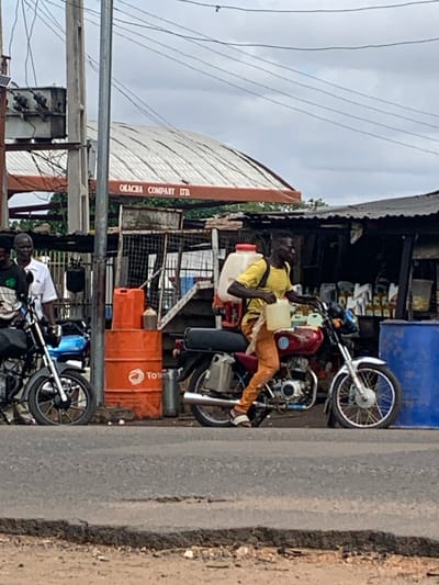 This image captures a street scene in Shendam, Nigeria, under an overcast sky. In the foreground, an asphalt road displays visible wear and an uneven edge. On the right side of the road, a dark-skinned man, wearing a yellow t-shirt, orange trousers, and white-striped sandals, is positioned on a red motorcycle with chrome accents and spoke wheels. He carries a white backpack-style sprayer and holds a translucent plastic container with a handle in his right hand. He is facing left, appearing to be in motion or preparing to move.
Behind the man on the motorcycle, an open-front stall with a dark corrugated metal roof displays multiple plastic bottles containing light-colored liquids, possibly oils or fuel. To the left of the red motorcycle, an orange metal drum with a partially visible "Total" logo is stacked with two smaller orange plastic jerry cans. A small, brown, conical container rests atop the stacked jerry cans. Further left, a dark-skinned man in a white short-sleeved collared shirt over a dark t-shirt stands beside a black motorcycle. His gaze is directed to the left. A partially visible arm and dark clothing suggest another individual further to the left.
In the background, a building with a distinctive curved, corrugated metal roof features a brown beam bearing the text "OKACHA COMPANY LTD." Utility infrastructure, including multiple poles (one metal, one concrete) and numerous overhead wires, crisscrosses the upper frame. A gray electrical transformer is visible on a pole structure on the far left. A large blue plastic barrel is partially visible on the far right. The overall scene depicts a bustling, informal roadside commercial area.
