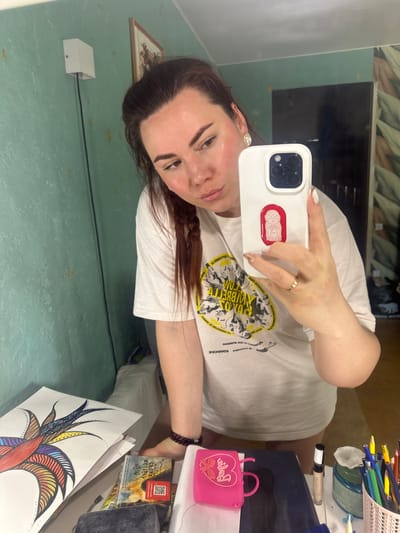 The image depicts a selfie taken by a woman with dark hair and fair skin. She is holding a white smartphone with a sticker of a baby in a red outline on the back. She is wearing a white T-shirt with a black and yellow graphic. A gold ring adorns the finger on her hand holding the phone.
The background includes a mirror reflecting a wall with a light fixture, and a desk cluttered with various items. On the desk, there are art supplies such as colored pencils in a container, a Barbie purse, a small grey box, and a drawing on paper with colorful line art.
Based on the OCR text "ЧЕРЁМУШКИ" visible on the box, the location may be in the Cheryomushki District of Moscow, Russia.