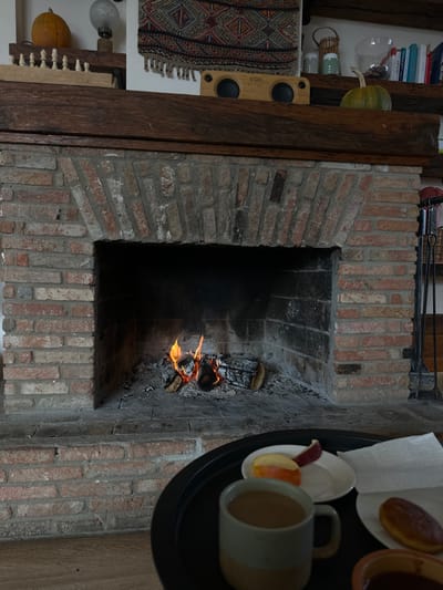 An interior scene in Sighnaghi, Georgia, depicts a lit brick fireplace with a tray of items in the foreground. The fireplace is constructed of reddish-brown bricks with an arched firebox containing burning logs, embers, and ash. Flames are visible within the firebox. To the right of the fireplace, a black metal stand holds fireplace tools. Above the fireplace, a thick, dark wooden mantelpiece supports various objects. On the mantel, from left to right, are a small orange pumpkin on a narrow wooden shelf, a decorative lamp with a frosted glass shade, a row of white chess pieces, a wooden "House of Marley" portable speaker with two circular grilles, and a green pumpkin. A patterned textile tapestry with geometric designs and fringe hangs on the wall above the speaker. Further right and higher, a small woven basket and several books with colorful spines are arranged on a wooden shelf. In the immediate foreground, a dark, round tray partially visible contains a two-toned mug filled with a light brown liquid, a white plate holding several slices of red and green apples, another white plate with a folded white napkin and a single pastry, and a partially visible small reddish-brown bowl. No individuals are present in the scene.