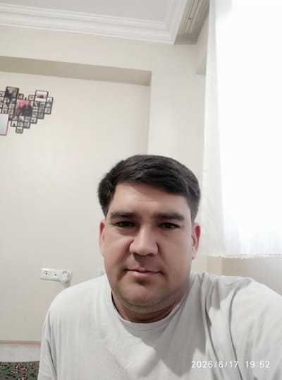 This is a close-up, vertically oriented selfie of a man, likely taken indoors. The man has dark hair and is wearing a white t-shirt. He is looking directly at the camera with a neutral expression. His face is partially obscured by the rotation of the image.
In the background, a cream-colored wall is visible. On the wall, there is an electrical outlet with a white plug inserted. Above and to the right, a photo frame collage is mounted, containing multiple small photographs. The scene appears to be a domestic setting, possibly a living room or bedroom. The lighting suggests it is daytime, but the exact time of day is not definitively clear from the image itself.
The bottom left corner of the image displays white text overlay, reading "2026/6/17 19:52", indicating the date and time the photo was taken. There are no obvious external location cues beyond the generic indoor setting. The overall mood is casual and personal, typical of a selfie.