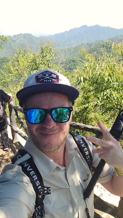 A man is taking a selfie outdoors with a scenic mountain and lush green vegetation background. He is wearing a tan button-up shirt, a grey baseball cap with a dark blue brim and a patch on the front that says "DARK SEAS", and dark sunglasses with blue mirrored lenses. The man has a light beard and is smiling. He is holding a black monopod or selfie stick in his right hand and is making a peace sign with his left hand. His backpack straps, with the word "CONVERSE" visible on them, are over his shoulders. In the background, there are steep, tree-covered mountains. The location is likely the Mutianyu section of the Great Wall of China in Huairou District, China, given the context of scenic mountain views and the presence of a wooden railing which is characteristic of tourist paths in that area. The sun is shining brightly, casting shadows.