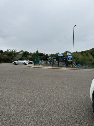 The image shows a playground enclosed by a green metal fence, located in Courtown, Ireland. The playground features blue and yellow climbing structures, slides, and a climbing wall. A few children and adults are visible within the fenced area. Outside the fence, to the left, are two silver cars parked in a paved area. A street lamp is visible in the background to the right of the playground. The sky is overcast, and the surrounding environment consists of trees and green foliage. The foreground is a grey asphalt surface, consistent with a road or parking lot.