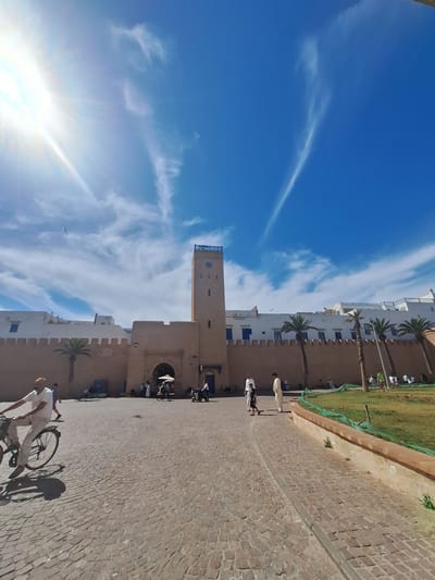 The image captures a bright, sunny day in the city of Essaouira, Morocco. The scene is dominated by a large, light-brown fortified wall with a prominent archway leading into the city. A tall, square clock tower, also light brown, stands to the right of the archway, topped with blue architectural details. White buildings with blue-trimmed windows line the top of the wall. Palm trees are scattered along the wall's base.
In the foreground, a man in a white shirt and light-colored pants rides a bicycle from left to right, casting a shadow on the cobblestone ground. Another person walks behind him. Further back, near the archway and the tower, several people are visible, some standing and others walking. One person is pushing a cart with a white umbrella.
To the right of the paved area, a patch of green grass is bordered by a low, reddish-brown wall. A green net is draped over this wall. People are scattered on the grassy area, some appearing to be children playing.
The sky above is a vibrant blue with wispy white clouds and the bright glare of the sun in the upper left corner, creating a lens flare effect. A bird is visible flying in the sky. The overall impression is a bustling public space in a historic coastal Moroccan city.