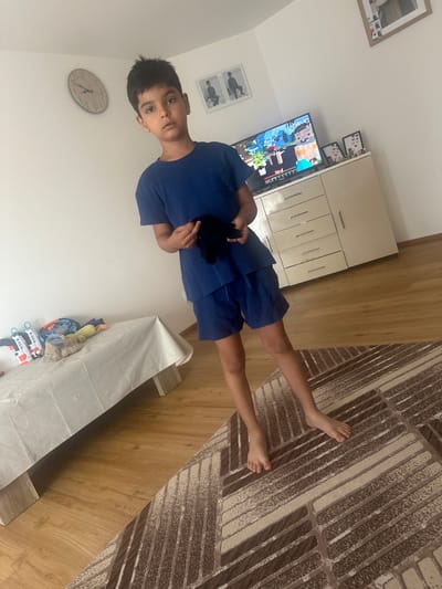 In an indoor setting, a young boy with dark hair and brown eyes, wearing a blue t-shirt and shorts, stands in the center of the frame. He holds a black object in his hands. The room features wooden flooring and a patterned rug. To the left, a bed is covered with a white cloth, and several toys are placed on it. A clock is mounted on the wall above the bed. To the right, a dresser with drawers and cabinets is visible, and a television screen displays a video game. Framed pictures are also present on the wall and on top of the dresser. The scene appears to be a child's bedroom or playroom. This image was captured in Mainburg, Germany.