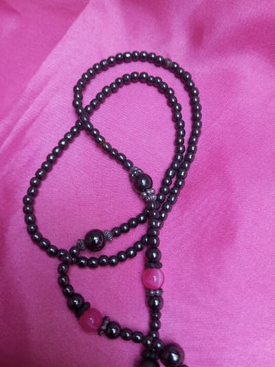 A rosary-style string of prayer beads is depicted resting on a vibrant pink fabric, likely satin or silk, which displays subtle folds and highlights. The beads are predominantly dark gray to black, spherical, and exhibit a metallic sheen. The strand incorporates several larger, spherical dark gray beads acting as dividers, along with two distinct larger, semi-translucent spherical pink beads. These larger beads, both dark and pink, are accented by small, silver-toned metallic spacers or caps. The beaded strand is arranged in multiple overlapping loops and curves across the fabric surface. No individuals are present. The scene is located in Qarabucaq qəsəbə, Azerbaijan.
