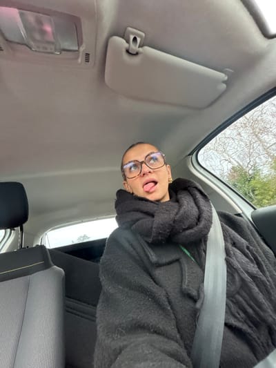 A person is seated in the front passenger seat of a vehicle, in what is identified as Saint-Cyprien, France. The individual, appearing female, has light-brown skin, dark hair pulled back, and wears large, round, tortoise-shell-framed eyeglasses. She is dressed in a thick, black, textured coat and a voluminous black knitted scarf. A dark grey seatbelt with a light grey stripe diagonally crosses her chest and lap. She is looking upwards and to the right, with her mouth open and her tongue extended. Her left ear displays a small gold hoop earring.

The vehicle's interior features a light grey ceiling, from which a central dome light fixture is visible, reflecting a faint pink hue. A grey sun visor is clipped up on the passenger side. Through the rear passenger window and front passenger window, a grey, overcast sky and bare tree branches are discernible, with some evergreen foliage also visible, suggesting an outdoor, possibly rural or suburban, setting. The upper portion of the rear seat headrest and the fabric of the rear seat are visible on the left side of the frame.