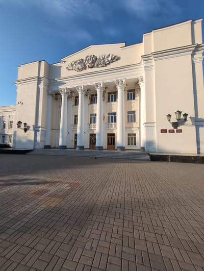 The image shows a light-colored building with neoclassical architectural elements, including tall white columns and decorative friezes. The building appears to be a public institution, possibly a theater or government building, given its grand facade. Several windows with blue-tinted panes are visible, and a wooden door is located centrally on the ground floor, accessed by a short flight of steps.

To the right of the building, a paved walkway made of rectangular bricks stretches into the distance. The bricks are laid in a herringbone pattern and some sections have a reddish-brown hue, possibly indicating a different type of paver or a shadow effect. The sun is casting long shadows across the walkway, suggesting it is either early morning or late afternoon. The sky is a clear, vibrant blue with a few small, dark specks that might be birds or insects.

On the wall of the building, above a textured band, there are three rectangular signs and a wall-mounted light fixture with three lanterns. The signs are too small to be read clearly, but they appear to be official notices. Another light fixture is visible lower down on the building's facade, near a security camera. The overall impression is one of a formal and stately public space. The image is oriented vertically, with the sky at the bottom and the ground at the top, implying it was taken with the camera held in a portrait orientation and then rotated. The scene is set in Tikhoretsk, Russia.