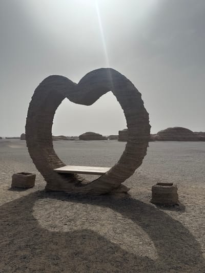 The image displays a large, heart-shaped structure made of a coarse, dark gray material, positioned on its side within a vast, arid landscape. The structure occupies the central-left area of the frame. Inside the lower curve of the heart, a light-colored wooden bench is visible.

A strong sun glare originates from the upper left corner, brightly illuminating the scene and casting a prominent, elongated shadow of the heart shape across the gravelly ground towards the right. Two smaller, cylindrical or square-shaped structures, made of similar material, are visible near the shadow and in the foreground.

The setting is a desolate desert environment, characterized by a flat, light brown-gray gravel terrain. In the mid-ground and background, distant, muted gray rock formations or hills stretch across the horizon beneath a bright, hazy sky. No people are visible.

The scene suggests a sunny day, likely late morning or early afternoon, given the intensity of the light and the distinct shadows. The heart shape, combined with the bench, implies a photo opportunity or a romantic landmark in this stark landscape, which is located in Dunhuang City, China. There is no visible text in the image.