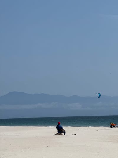 This image depicts a wide, light-colored sandy beach in La Cruz de Huanacaxtle, Mexico, under a clear, pale blue sky. A distant, hazy blue-grey mountain range with low-lying clouds forms the horizon above a dark blue ocean exhibiting moderate wave activity. In the midground, a lone individual wearing a red cap, blue shirt, and dark pants is seated on a dark, weathered log or tree stump, facing away from the camera towards the ocean and mountains. A dark shoulder bag is visible on the person. To the right, above the ocean, a blue and white kiteboarding kite is airborne. Further right, closer to the water's edge, an orange wheelbarrow containing several yellow, round objects is present on the sand.