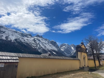 The image shows a street view in Sappada, Italy, featuring a low yellow wall with a wooden roof. To the left is a wooden gate with a peaked roof. Behind the wall is a mountainous landscape with snow-capped peaks and a dark green forest. The sky is blue with scattered white clouds. To the right of the wall, there is an ornate gate structure with a cross on top and the inscription "RESURRECTIO". Bare trees are visible alongside the wall and gate. A gray paved road is visible in the foreground. The overall impression is a landscape combining architectural and natural elements under a partly cloudy sky.