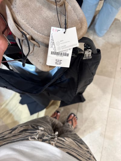 The image shows a close-up view of a person's legs and feet, wearing striped pants and flip-flops, in what appears to be a retail store. The person is standing on a tiled floor. In the background, a beige-colored hat with a black string is hanging from a rack, attached to a price tag. The price tag displays information in multiple languages and includes the prices US $59.90 and UK £29.99. The store appears to be a Zara branch in Dartford, United Kingdom, suggested by the presence of a Zara logo visible on a tag in the background. The lighting suggests an indoor environment.