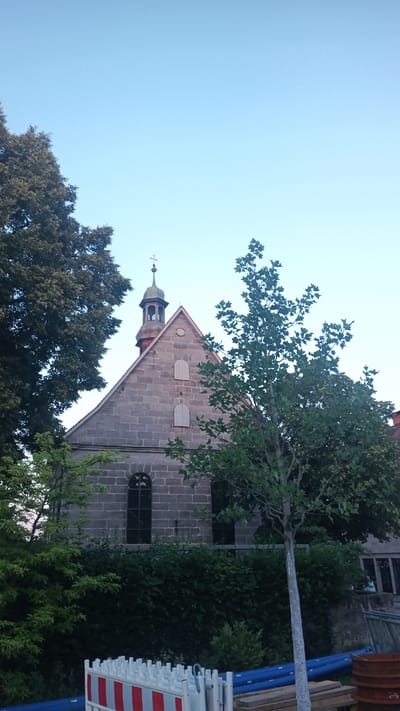 The image depicts a stone building with a steeple and a cross, likely a church, situated in Altdorf bei Nürnberg, Germany. The building is made of roughly cut stone blocks, and its roof is steep. A small bell tower with a green copper roof is visible on top of the main roof, topped by a cross. The building has a large arched window on the ground floor and two smaller, rectangular shuttered windows on the upper level. The scene is framed by lush green trees and bushes, some of which are significantly larger and older than others. In the foreground, there are construction barriers, including red and white striped barriers and blue pipes, suggesting ongoing work or maintenance in the area.

The time of day appears to be late afternoon or early evening, given the soft, diffused light and the clear, pale blue sky. The weather seems calm and pleasant. The surrounding vegetation is vibrant and full, indicating a period of growth, likely summer. The overall atmosphere is serene, with the solid stone structure of the church contrasting with the organic, living elements of the trees and foliage. There is no visible text in the image. The scene suggests a quiet, possibly historic, setting.