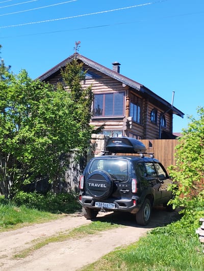 A black Lada Niva Travel car, with a roof box and a spare tire mounted on the back, is parked in front of a log cabin house. The car's license plate reads "P381EE 716 RUS". The house has a decorative roof trim and a weather vane on top. The setting is a rural area, with dirt roads and trees surrounding the house. The sky is clear and blue. The location is presumably in or near Икское Устье, Russia, based on the license plate information.