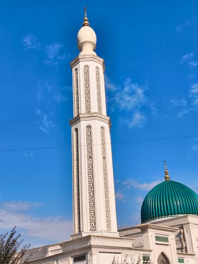 A low-angle, full outdoor shot captures the exterior of a mosque under a bright blue sky with scattered white clouds. The main subject is a tall, white minaret with ornate decorative carvings that rises on the left side of the frame. The minaret is topped with a golden finial featuring a crescent moon. To the right of the minaret, a vibrant teal-green dome of the mosque is visible, also adorned with a golden crescent moon at its peak. The mosque structure itself appears to be white with intricate architectural details. A single thin black wire is stretched horizontally across the middle of the sky. In the bottom left corner, the branches of a dark-leaved tree are partially visible, suggesting a natural element near the building. The scene depicts a religious structure in an urban or semi-urban setting, likely Buz, Uzbekistan.