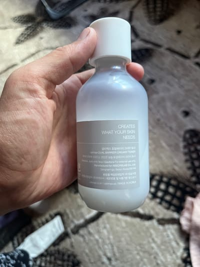 A person's hand holds a white bottle of skincare product. The bottle has a light grey label with white text. The text on the label is partially visible and includes "CREATES WHAT YOUR SKIN NEEDS" in English, and Korean text which appears to be product information. Some of the Korean text includes "셀리맥스 듀얼배리어 크리미 토너" and "celimax DUAL BARRIER CREAMY TONER". Additional details include "Manufactured for ABSORBLAB Co., Ltd." and "MADE IN KOREA". The background is a blurred pattern of fabric. The lighting suggests it is daytime, and the setting appears to be indoors.
