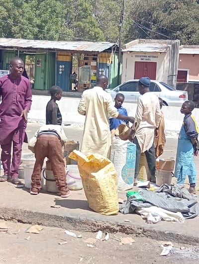This image depicts an outdoor commercial or market scene in Zango, Nigeria, under bright daylight. In the foreground, multiple individuals are gathered near various goods. An adult male in a purple garment stands on the far left, looking towards the left. To his right, a child wearing brown trousers and a light top is bent over, facing away from the viewer, near several white plastic buckets.

In the center, two adult males, both in light-colored tunics, are interacting with children and large sacks. One adult male, facing right, appears to be handing something to a child in a blue shirt. The other adult male, wearing a dark cap and facing left, is near a large white sack labeled "TRS PREMIUM WHEAT FLOUR." Another child in a light blue garment is partially visible on the far right, looking left. A large yellow sack stands upright on the ground near a pile of assorted fabrics. The ground in the immediate foreground is unpaved dirt with scattered debris, including cardboard pieces.

In the mid-ground, a silver sedan car is parked. Behind the individuals, multiple structures are visible. To the left, a low-rise building constructed from repurposed container-like units, painted green and blue with yellow accents, features a corrugated metal roof. To the right, a smaller, light-colored concrete building with a red door or shutter bears the inscription "EDUCATIONAL MATERIALS AND SHOES" above its entrance. Green trees are present in the distant background.