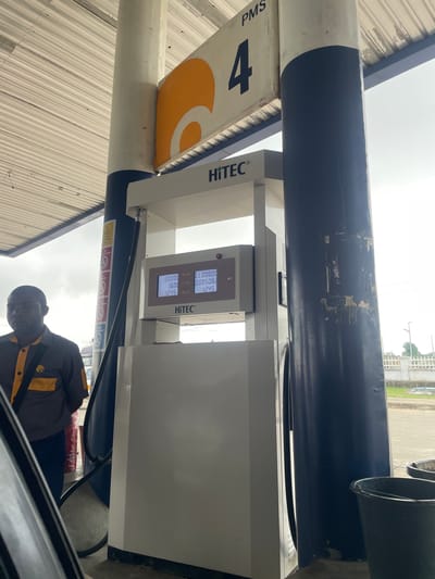 An outdoor scene at a gas station, likely in Karmajiji, Nigeria, features a fuel dispenser with a digital display and a male attendant standing to the left. The fuel dispenser, labeled "HiTEC," shows transaction details including amounts in NAIRA, LTRF, and PRICE. The attendant, wearing a gray and yellow uniform with a black strap across his chest, is looking downwards. The dispenser is situated under a covered area with a ribbed metal roof. Behind the dispenser, a large, dark blue pillar supports the roof structure. In the background, a fence and some vegetation are visible under an overcast sky. Several buckets are placed on the ground near the dispenser.