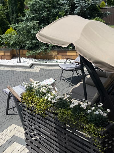 A shaded outdoor seating area features a tan swing seat with dark gray metal framing and cushions. White flowers with green foliage are planted in a dark gray planter box along the railing. The ground is paved with gray and beige interlocking bricks. To the left of the swing, a pine tree with blue-green needles is visible, along with other greenery. The scene suggests a pleasant day outdoors, possibly in a garden or patio setting. The lighting indicates it's daytime, with shadows cast by the swing and railing. The location is identified as Koroviia, Ukraine. There is no visible text in the image.