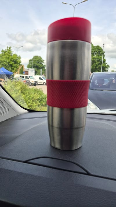 A stainless steel travel mug with a red lid and a red textured grip is placed on the dashboard of a vehicle. The dashboard is dark gray and textured. Through the windshield, a cloudy sky with some blue patches is visible, along with trees, streetlights, and several parked vehicles. The scene appears to be in an outdoor parking lot. This image was taken in Preiļi, Latvia.