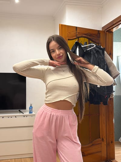 A young woman poses indoors, holding a smartphone near her head. She wears a beige, long-sleeved shirt and pink sweatpants. Behind her is a wooden cabinet with clothing hanging on it, and a TV mounted on a white wall. The location is likely a residential room. There is no evidence to conclude it's Kovel, Ukraine.
