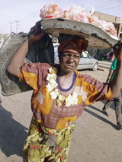 The image displays an outdoor street scene in Kwekwe, Zimbabwe, under bright daylight. The central subject is an elderly woman with dark skin, depicted from the waist up. She wears a red and brown patterned headwrap, a brown and orange geometric-patterned long-sleeved shirt, a blue beaded necklace, and a second necklace of light-colored, possibly artificial, flowers. Her lower attire consists of green and red floral-patterned trousers. She is actively balancing a large, white, rectangular metal tray on her head with her right hand. This tray holds several clear plastic bags, each containing multiple reddish-orange spherical items, consistent with produce like tomatoes or oranges. Her left arm is raised, supporting a dark, possibly woven fabric bag or garment over her left shoulder. The woman's facial expression shows a furrowed brow and slightly open mouth. In the immediate background to the right, a person wearing dark trousers and a green shirt is partially visible, positioned behind and to the side of the central figure. Further in the background, a light-colored sedan car is visible, alongside utility poles with overhead lines against a clear, bright sky. The ground surface is asphalt pavement.