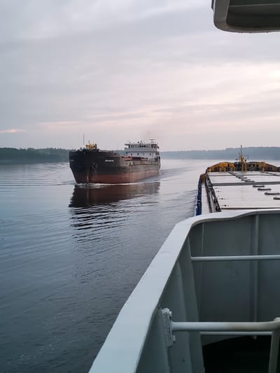 The image captures a calm river scene with two barges. In the foreground, the deck of one barge is visible, showing yellow containers and a white railing. In the mid-ground, a larger barge, painted red and black, is heading towards the viewer, leaving a wake in the water. Its hull bears Cyrillic lettering which appears to read "ВОЛГО-БАЛТ 113". The background shows a tree-lined shore under an overcast sky. The location is identified as the city of Ratibor, Russia.