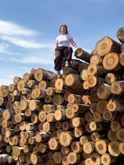 The image shows a woman holding onto a stack of freshly cut logs under a blue sky. The logs appear to be birch, identified by their white bark, and they are neatly stacked, with the cut ends prominently visible. The woman is wearing a white shirt and brown pants. The sky shows faint clouds. The image orientation is rotated, giving the impression of the logs hanging from the sky and the woman holding onto them. The specific location in the city of Ural'skoe, Russia, is not discernible from the image alone.