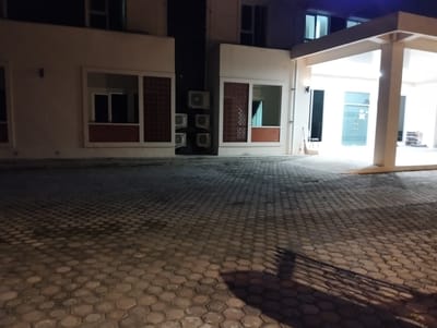 This image shows an exterior view of a modern building at night, possibly a hotel or a commercial establishment, in Palohgadeng, Indonesia. The foreground consists of a hexagonal-patterned paved driveway, casting long shadows from an unseen light source. The building features a prominent covered walkway with large pillars and a white ceiling. To the left, there are sections of the building with windows featuring decorative latticework and what appear to be air conditioning units mounted externally. The windows are dark, indicating the interior is not brightly lit. The overall lighting suggests nighttime, with artificial lights illuminating the walkway and parts of the building exterior, contrasting with the darker paved area.