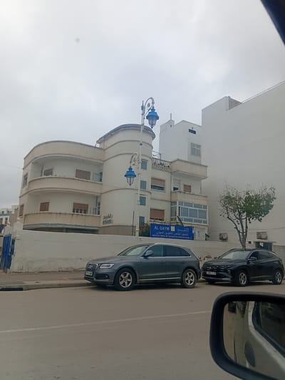 This media file captures a street scene in Martil, Morocco, on an overcast day. The photograph appears to have been taken from inside a vehicle, with a portion of a car's side mirror visible in the upper right.
In the midground, a paved road stretches, along which two cars are parked parallel to the curb. The first car is a dark grey sedan, facing away from the viewer, with a visible license plate reading "7711NBB". Behind it is a black sedan, whose license plate displays "14680 111 75".
To the left of the street stands a distinctive multi-story white building featuring a curved facade and balconies. Several ornate blue streetlights are attached to its exterior. Further down the street, another white building is visible, fronted by a small tree with green foliage. This building features a prominent blue sign that reads "AL QAYM" in Latin script and "القيم" in Arabic, followed by "المعهد الخاص للتكوين المهني" and "INSTITUT PRIVE DE FORMATION PROFESSIONNELLE," indicating a private professional training institute.
The sky is a uniform light grey, suggesting an overcast or cloudy day. No people are visible, and no specific activity is taking place; the scene appears tranquil.