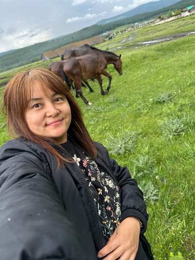 A woman with brown hair, wearing a black jacket over a floral shirt, takes a selfie in a grassy field. In the background, two horses graze in a muddy field, with a village and rolling hills in the distance. The sky is overcast. The scene is likely in Abzakovo, Russia, a popular resort town known for its natural beauty and horse riding opportunities.