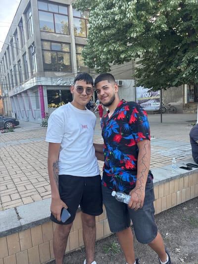Two men are standing together on a brick sidewalk next to a street. One man wears a white t-shirt and black shorts with sunglasses, holding a phone in his hand. The other wears a floral patterned shirt and gray shorts and holds a bottle of water. A building with multiple windows is visible in the background, along with a tree with dense foliage. A bicycle is parked near the building, and a parked car is partially visible further back. The location appears to be Hayredin, Bulgaria.