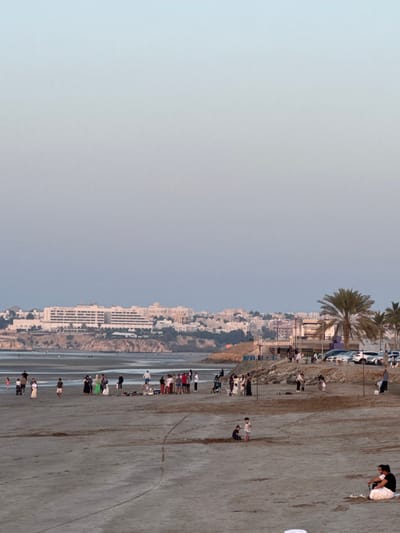 The image depicts a panoramic view of Qurum Beach in Bawshar Province, Muscat, Oman, during twilight hours. The foreground consists of a broad expanse of light-brown sandy beach exhibiting faint linear indentations. In the lower right, an individual is seated directly on the sand, wearing dark attire and a white garment around the lower torso.
Approximately twenty to thirty individuals are scattered across the mid-ground beach. Several small groups are engaged in leisure activities, with some utilizing blankets on the sand. Two children are visible centrally, one standing and one kneeling, appearing to play. To the right, a paved parking area adjacent to a rocky embankment holds multiple parked vehicles, predominantly white, with palm trees rising behind them. Individuals are observed walking, standing, sitting, and engaging in casual recreation.
The background presents a coastal cityscape across a body of water. The dominant structure is a large, multi-story white hotel complex, identified as 'CROWNE PLAZA', situated on an elevated landmass. Numerous other white and light-colored buildings comprise the urban sprawl extending along the coastline and inland hills. The sky displays a clear gradient from light blue overhead to a softer, grayish-purple hue near the horizon.