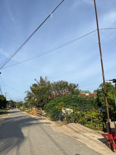 This is a street-level, eye-level, medium shot of a paved road in Landuh, Indonesia, with a clear blue sky overhead. The road stretches into the distance, lined with various trees, plants, and buildings. On the right side of the road, a rusty metal pole stands vertically, supporting overhead wires. A red wheelbarrow is partially visible at the bottom right corner of the image. In the mid-ground, there's a dense collection of potted plants and a small pile of sand. Further down the road, several houses with corrugated metal roofs can be seen, interspersed with lush greenery and palm trees. A single person on a motorbike is visible on the road in the distance. The overall impression is of a rural or semi-rural residential area under bright daylight.