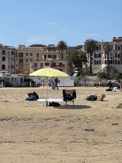 The image depicts a sandy beach in Anzio, Italy. Parallel to the beach is a line of multi-story buildings with architectural styles ranging from older structures with arched windows to more modern buildings with balconies and green shutters. A few palm trees are interspersed among the buildings.
On the beach, there are people relaxing, walking, and interacting. A yellow and white striped umbrella stands near the building. Beach chairs, towels, and bags are scattered around, indicating people are spending time on the beach. The sand shows signs of foot traffic and tire tracks.