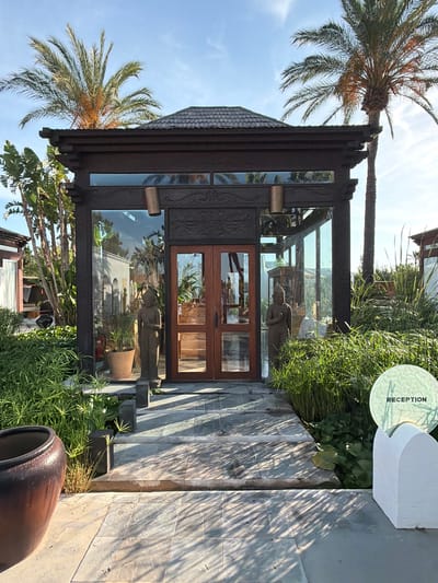 The image shows the entrance to a building in Sant Joan de Labritja, Spain. The entrance is a dark wood structure with glass walls and a dark tiled roof. Two large wooden doors are at the center of the entrance. In front of the entrance, there are two stone statues of figures with their hands clasped. Lush green plants and palm trees surround the entrance. A stone pathway leads up to the doors, and to the right, there is a white, arched structure with a round, textured glass sign that reads "RECEPTION". The sun is shining, casting shadows on the pathway and the surrounding foliage.