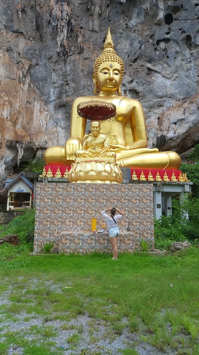 A large golden Buddha statue, partially set into a rocky hillside, dominates the scene. The Buddha is holding a round object. To the right of the statue, a young woman with a ponytail is standing on a patterned tile surface and appears to be looking at something on the ground. A yellow object is also on the tile surface. The environment includes green grass, small building in background, and other foliage. The location is Thap Put, Thailand.
