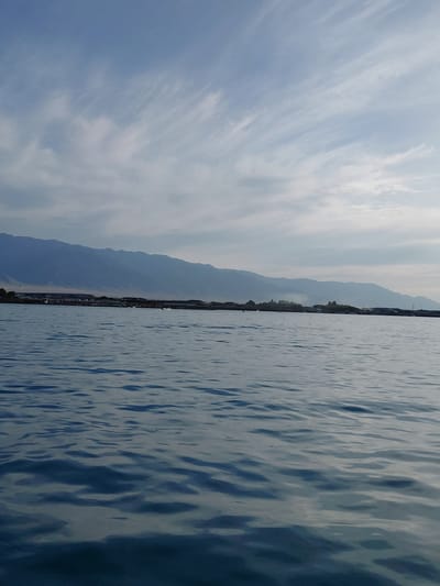 This is a landscape photograph taken from the water, looking towards a shoreline with mountains in the background. The image is oriented vertically, with the water occupying the right two-thirds of the frame and the sky and land occupying the left third.
The water is choppy, with ripples and reflections creating a textured surface. The color of the water transitions from a lighter blue in the foreground to a darker blue towards the center of the frame. The shoreline is lined with buildings, suggesting a town or city. Beyond the buildings, a range of mountains is visible, appearing hazy and faded due to distance. The sky is mostly clear with some wispy white clouds that stretch across the frame.
The time of day appears to be late afternoon or early evening, given the soft light and the angle of the sun which is not directly visible. The weather seems clear and calm, with no signs of rain or strong winds. The location is Akshi, Kazakhstan, a place known for its natural beauty and proximity to mountains and bodies of water.
No people or specific objects are discernible in the image, emphasizing the natural landscape. The overall emotion conveyed by the image is one of tranquility and expansive natural beauty. No text is visible in the image.