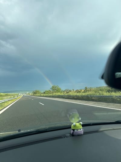 A view from inside a vehicle traveling along a highway. A rainbow is visible in the sky to the left. A small, crocheted owl is attached to the passenger-side window. The location is likely near Mlada Gvardia, Bulgaria. The environment consists of an open highway with greenery on both sides.