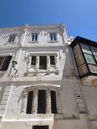 The image captures a close-up, low-angle view of the facades of two adjacent buildings under a clear blue sky. The building on the left is a white, stucco structure with ornate architectural details. It features several windows, some with dark shutters, and decorative moldings. The stucco is weathered and peeling in places, revealing an older layer underneath. A black, angled street lamp is mounted on this facade. The building on the right is darker, with a prominent bay window that extends outwards. This bay window is made of dark wood or metal framing and contains multiple panes of glass, some with curtains visible. The facade of this building appears to be a dark color, possibly black or very dark brown, with ornate yellow decorative elements visible beneath the bay window. The architectural styles suggest a historical European city. This view is located on a street in the historic center of Maó, Spain.