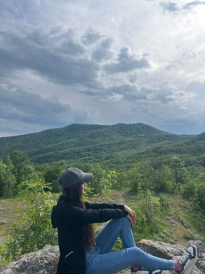 A medium, angled shot captures a person seated on a rock outcrop, with their legs outstretched and feet pointing towards the top right of the frame. The person wears a black jacket, blue jeans, and athletic shoes with light soles and dark accents. A gray baseball cap covers their head. Their hands are resting on their knees.
Behind the person, a lush, green, tree-covered hillside fills a significant portion of the frame, rising up and to the left. To the left of the hillside, the sky is visible, with overcast and cloudy weather. The scene is presumably located in the vicinity of Kamchia, Bulgaria, given the instructions. The environment appears natural, showing the elements of a forest and a rocky terrain. The overall impression is of a solitary person enjoying a view in a natural setting.