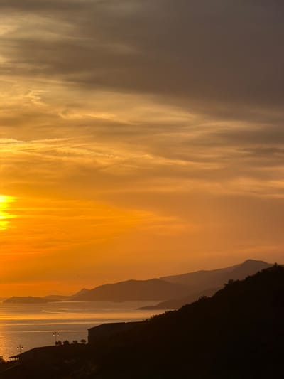 This image captures a serene sunset over the Adriatic Sea, viewed from Krimovice, Montenegro. The sky is a gradient of warm oranges and yellows, with wispy clouds stretching across the horizon. Silhouetted against the vibrant sky are layers of distant mountains, their shapes softened by the fading light. In the foreground, a dark, hilly landscape is visible, with the faint outlines of buildings and streetlights suggesting human habitation. The calm sea reflects the golden hues of the sunset, creating a shimmering path of light across its surface. The overall scene is tranquil and picturesque, highlighting the natural beauty of the Montenegrin coastline at dusk.