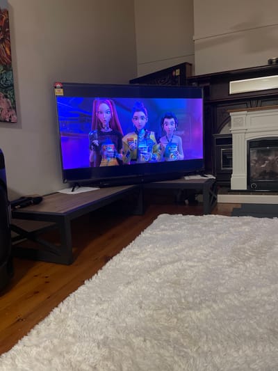 The image displays a television screen showing an animated scene with three young women. They appear to be characters from a children's show or movie, with stylized features and colorful outfits. One character on the left has long, dark red hair and is wearing a black outfit adorned with studs. The middle character has dark hair styled in a bob and is dressed in a yellow and blue outfit, holding a can. The character on the right has lighter hair and is also wearing a yellow and blue outfit, holding a similar can. The background on the screen is a vibrant purple, suggesting an indoor or nighttime setting.

The television is placed on a dark wooden media console with black metal accents. The console has an open shelf where some black objects are visible, possibly electronic devices or remotes. To the right of the console, a large, fluffy white rug covers a portion of the wooden floor. In the background, a fireplace with a dark mantle and white trim is partially visible. The overall scene suggests a casual, indoor entertainment setting, likely a living room. The image is taken from a slightly elevated angle, looking down at the television and the surrounding area. The lighting appears to be dim, typical of an evening or a room with the lights turned down for watching television. There is no indication of time of day or weather conditions. The emotions conveyed by the characters on screen are neutral or slightly upbeat.

Visible text on the screen includes "ZOEY" on one of the cans held by a character, and potentially other text on the cans and clothing that is not clearly legible. There is also a small yellow and red sticker on the bottom right corner of the television, which appears to be an energy rating label, commonly found on electronics in Australia. The location context of Bendigo, Australia is provided.