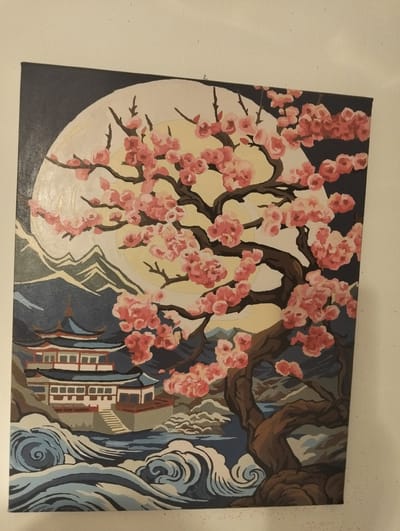 This is a painting of a cherry blossom tree in front of a full moon, with a traditional East Asian-style building in the background. The building is multi-tiered with a dark blue roof and white walls, and it is situated near a body of water with stylized waves. Mountains are visible behind the building. The cherry blossom tree has a dark brown trunk and branches, adorned with numerous pink blossoms. The moon is a pale yellow circle against a dark gray sky. The painting is rendered in a flat, stylized manner with bold outlines. The image is a photograph of the painting hanging on a plain white wall. There is no indication of any specific location in the city of Қараөткел, Kazakhstan.