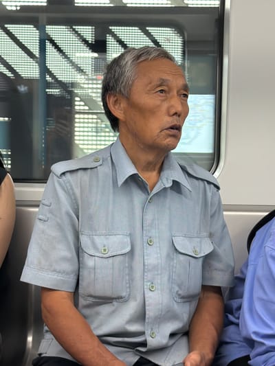 The image shows an elderly Asian male seated in a public transport vehicle, likely a subway or train car, in Tiaodeng, China. He has gray hair, a tanned complexion, and visible facial wrinkles. He is wearing a light blue, short-sleeved, collared shirt with thin vertical stripes, two buttoned chest pockets, and buttoned shoulder epaulets. The buttons on his shirt are light green or beige. Two small fabric loops are affixed to the upper left sleeve. His mouth is slightly open, and his gaze is directed towards the upper right quadrant of the frame. To his immediate left, the upper arm and shoulder of another individual wearing a dark garment are partially visible. To his right, a segment of a third person clad in a plain light blue shirt is present. Behind the primary subject, a window displays a blurred map or diagram along with a prominent white grid pattern, possibly from internal lighting or external structures. The interior walls of the vehicle are white, and the visible seating appears to be metallic.